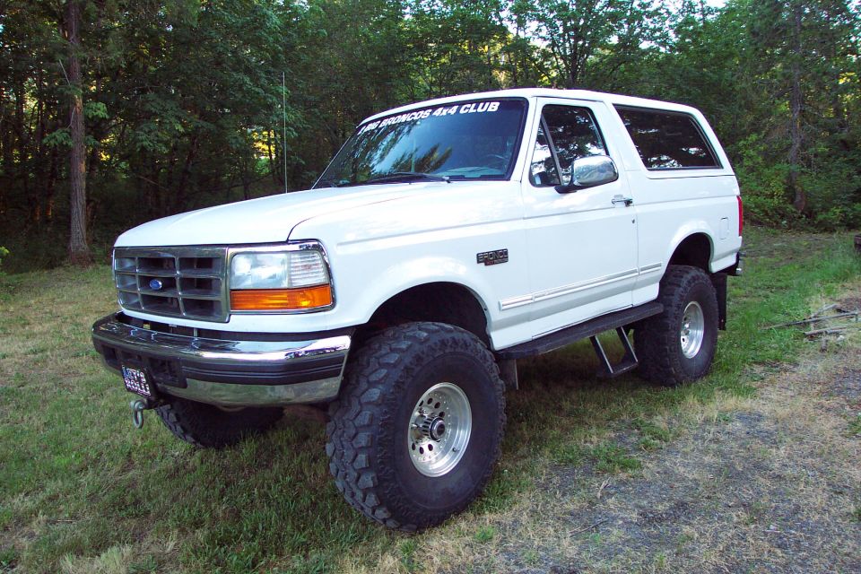 White lifted Ford Bronco on large mud tires parked at edge of woods