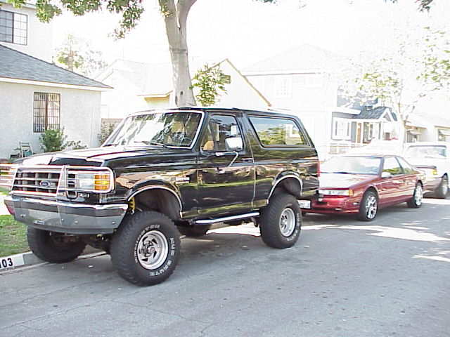 Black lifted Ford Bronco on a residential street with red coupe parked behind