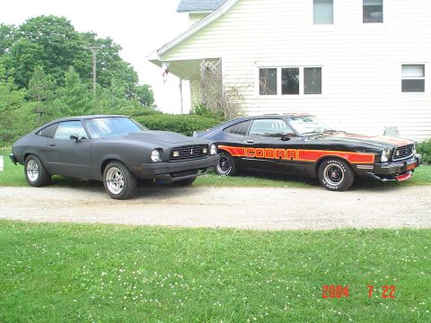1978 Ford Mustang II - Two 1978 Ford Mustang IIs, one matte black with a hood scoop and one black with orange Cobra stripes, sit parked in front of a house.