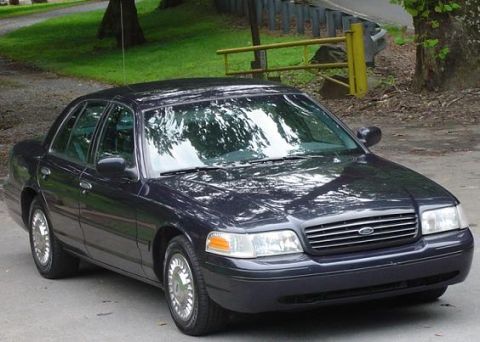 1999 Ford Crown Victoria - This dark sedan is parked outdoors with trees and a fence visible behind it.