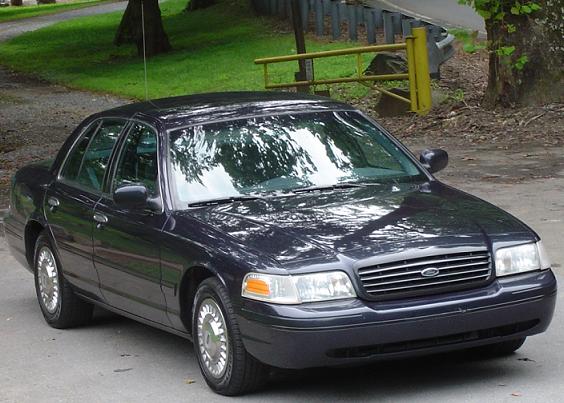 Dark sedan parked on a paved surface with trees and a yellow fence in the background