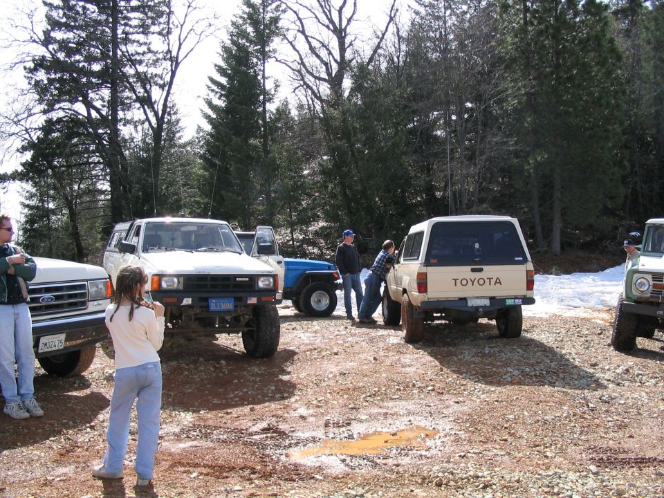 Several off-road vehicles parked on a dirt lot with trees in the background. A white Toyota SUV is visible