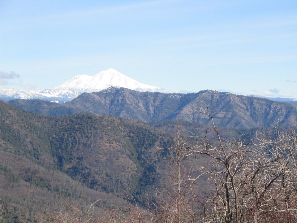 Mountain landscape with snow-capped peak in background and bare trees in foreground. Watermark reads SUPERMOTORS