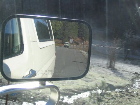 1989 Ford Bronco - Reflection in the side mirror of a white van shows a green SUV driving on a road with trees and a fence.