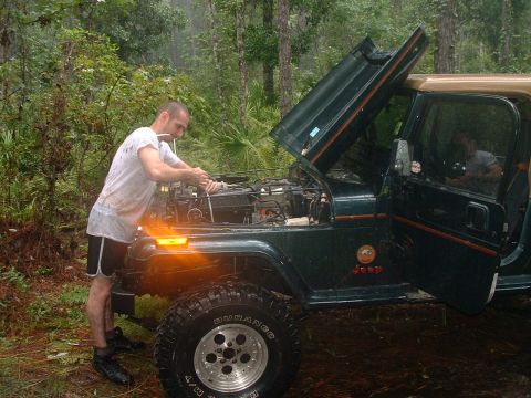 1995 Ford Bronco - A man works on the engine of a green Jeep Wrangler in a wooded area.