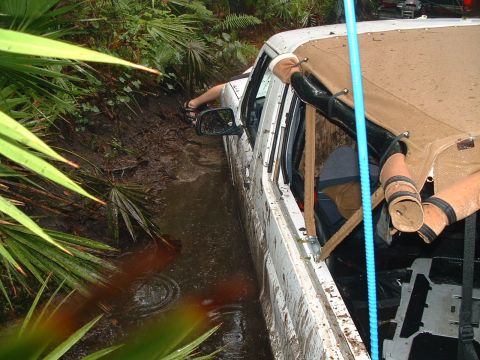 1995 Ford Bronco - A white 1995 Ford Bronco is stuck in a muddy, overgrown area, with its side visible and vegetation surrounding it.