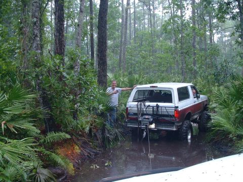 1995 Ford Bronco - A man stands next to a red and white Ford Bronco navigating a muddy trail through a dense forest.