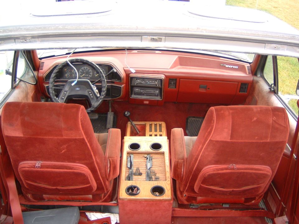 1987 Ford Bronco interior with red velour seats, woodgrain center console with cup holders, and dashboard with radio and gauges