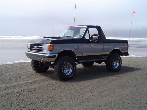 1989 Ford Bronco - This lifted 1989 Ford Bronco is parked on a beach, with the ocean in the background and an orange flag on a pole.