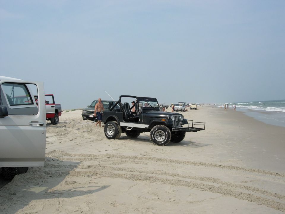 Black Jeep CJ-7 parked on a beach with a silver Ford truck in the foreground and other vehicles and people in the background near the ocean