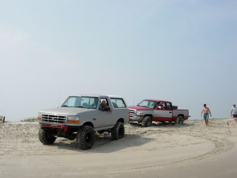 1995 Ford Bronco - A gray Ford Bronco and a red and silver pickup truck drive on a sandy beach under a clear sky.