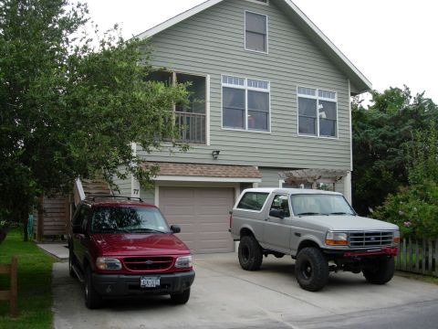 1995 Ford Bronco - A gray 1995 Ford Bronco with a white top and large off-road tires is parked in front of a house, alongside a red SUV.