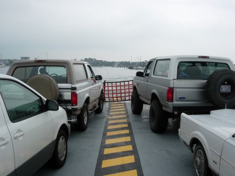 1995 Ford Bronco - Two Ford Broncos are parked on a ferry, with a white car in front and a body of water in the background.