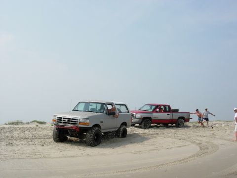 1995 Ford Bronco - A silver 1995 Ford Bronco is being towed by a red pickup truck on a sandy beach with people nearby.