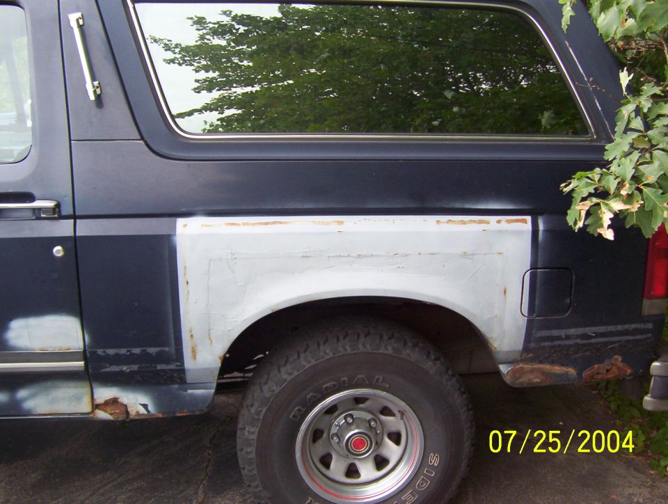 Dark blue Ford Bronco with rust and body filler on the rear quarter panel, seen from the side