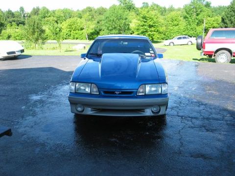 1990 Ford Mustang - A blue Ford Mustang with a prominent hood scoop is shown from the front, with other vehicles in the background.