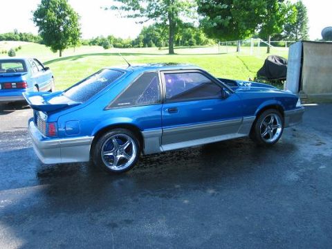 1990 Ford Mustang - A blue 1990 Ford Mustang coupe is parked on wet asphalt, with another blue car visible in the background.