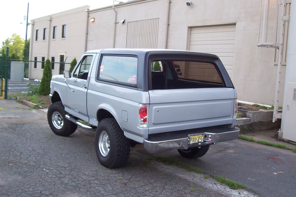 Lifted silver Ford Bronco with black top, rear 3/4 by an industrial building