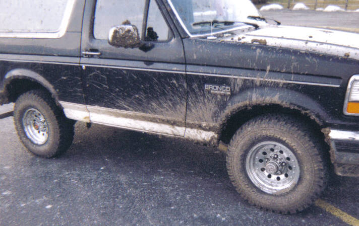 Mud-splattered black Ford Bronco SUV with chrome wheels and off-road tires, passenger side view in a parking lot