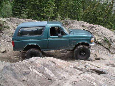 1996 Ford Bronco - A green 1996 Ford Bronco navigates a rocky off-road trail with trees in the background.