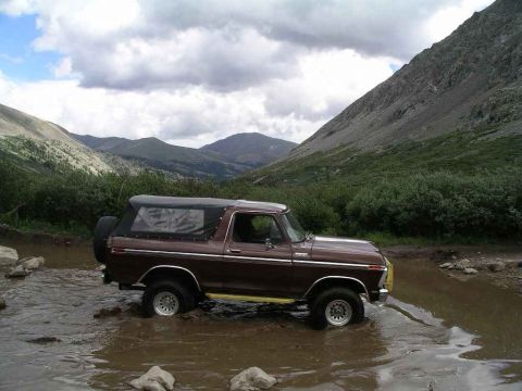 1996 Ford Bronco - Ford Bronco navigates a rocky stream bed surrounded by green hills and cloudy skies.
