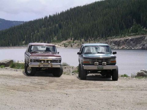 1996 Ford Bronco - Two Ford Broncos, one teal and one maroon, are parked near a lake with a forested mountain in the background.