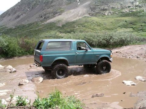 1996 Ford Bronco - A green Ford Bronco navigates a muddy obstacle on a dirt road with a mountainous backdrop.