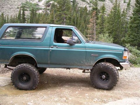 1996 Ford Bronco - A green Ford Bronco is parked on a dirt road with a wooded mountain landscape behind it.