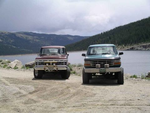 1996 Ford Bronco - A teal 1996 Ford Bronco is parked next to an older model Ford truck near a lake.