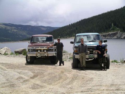 1996 Ford Bronco - Two Ford trucks are parked near a large body of water with a mountainous, forested backdrop under a cloudy sky.