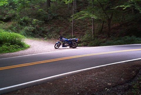1985 Ford LTD Crown Victoria - A blue motorcycle is parked on the shoulder of a road that winds through a forest.