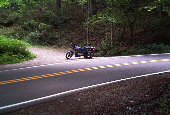 Blue motorcycle parked on the side of a two-lane road in a wooded area