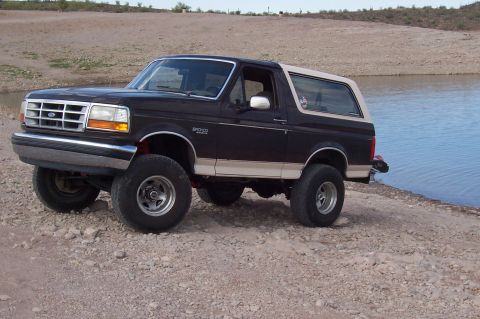 1992 Ford Bronco - Lifted dark blue Ford Bronco full-size SUV with tan two-tone lower body and tan hardtop, photographed from a 3/4 front angle on a rocky sho…