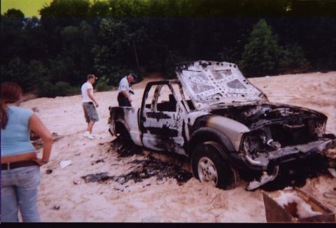 1989 Ford Bronco - A severely damaged pickup truck sits partially buried in sand, its hood open and interior scorched.