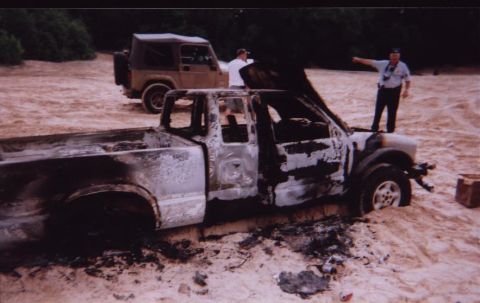 1989 Ford Bronco - A severely damaged pickup truck sits in sand next to a Jeep, with two men nearby.