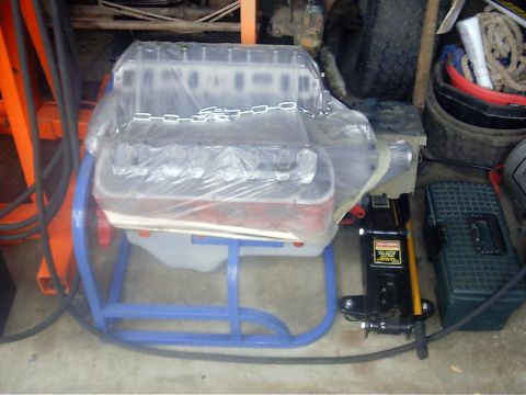 1990 Ford Bronco - An engine sits on a stand, covered in protective plastic, with a car jack nearby.