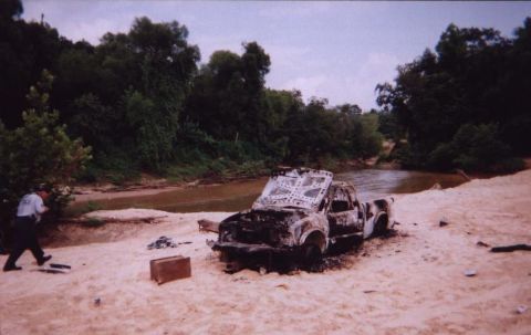 1989 Ford Bronco - A severely damaged pickup truck sits on a sandy shore next to a river, with a person in uniform observing the scene.