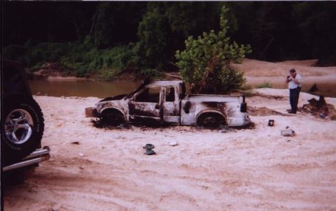 1989 Ford Bronco - A severely damaged pickup truck sits on a sandy bank near a body of water, with a person standing nearby.