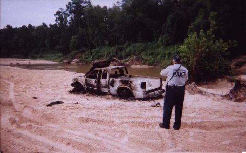 1989 Ford Bronco - A police officer surveys a heavily damaged pickup truck abandoned on a sandy riverbank.
