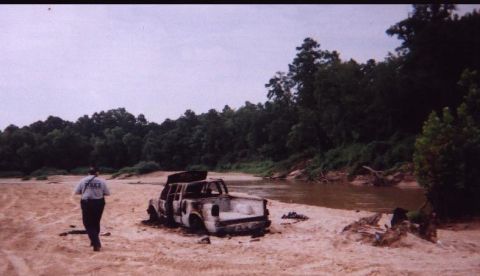 1989 Ford Bronco - A police officer surveys a burnt-out 1989 Ford Bronco abandoned on a sandy riverbank.