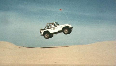 1990 Ford Bronco - A white Jeep Wrangler is captured mid-air, soaring over a sandy landscape with a small red flag trailing behind it.