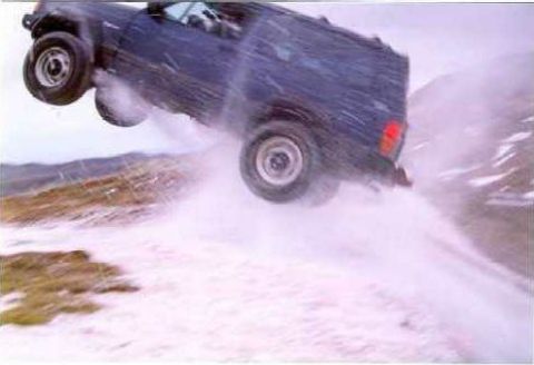 1990 Ford Bronco - SUV airborne during a jump over snowy terrain, creating a spray of snow.