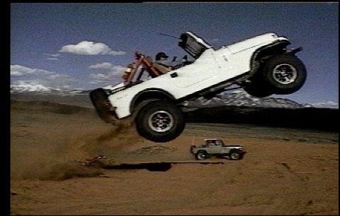 1990 Ford Bronco - A white off-road vehicle is captured mid-air during a jump over a sandy landscape.