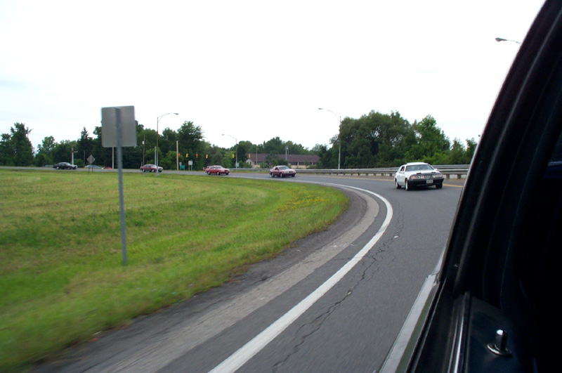 White coupe ahead on a curving ramp with dark red cars across the median, shot from a car