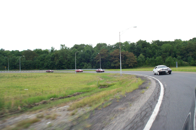 White full-size sedan ahead on a curving ramp with three dark red cars across the median