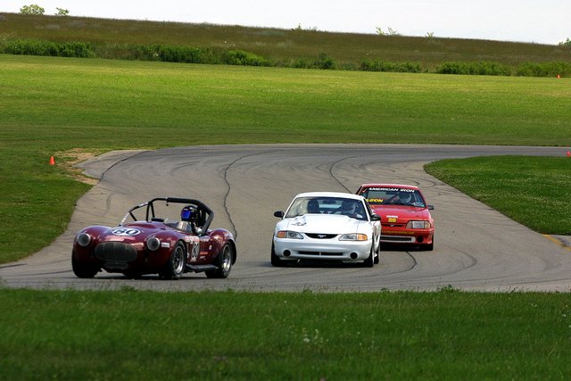 Dark AC Cobra-style roadster number 80 leading a white Mustang and red Fox-body through a turn