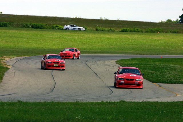Three red Mustangs cornering on a road course with a white car cresting the hill behind