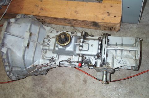 1995 Ford Bronco - A cast-aluminum manual transmission lying on a concrete garage floor next to a wooden beam, viewed from above with the bellhousing at left,…