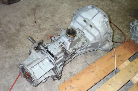 1995 Ford Bronco - Aluminum manual transmission lying on a concrete floor beside a wooden beam, viewed from the tailshaft end with the splined output shaft an…