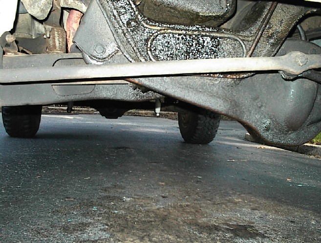 Underside view of a Bronco's front solid axle, steering linkage, and engine bottom over wet pavement